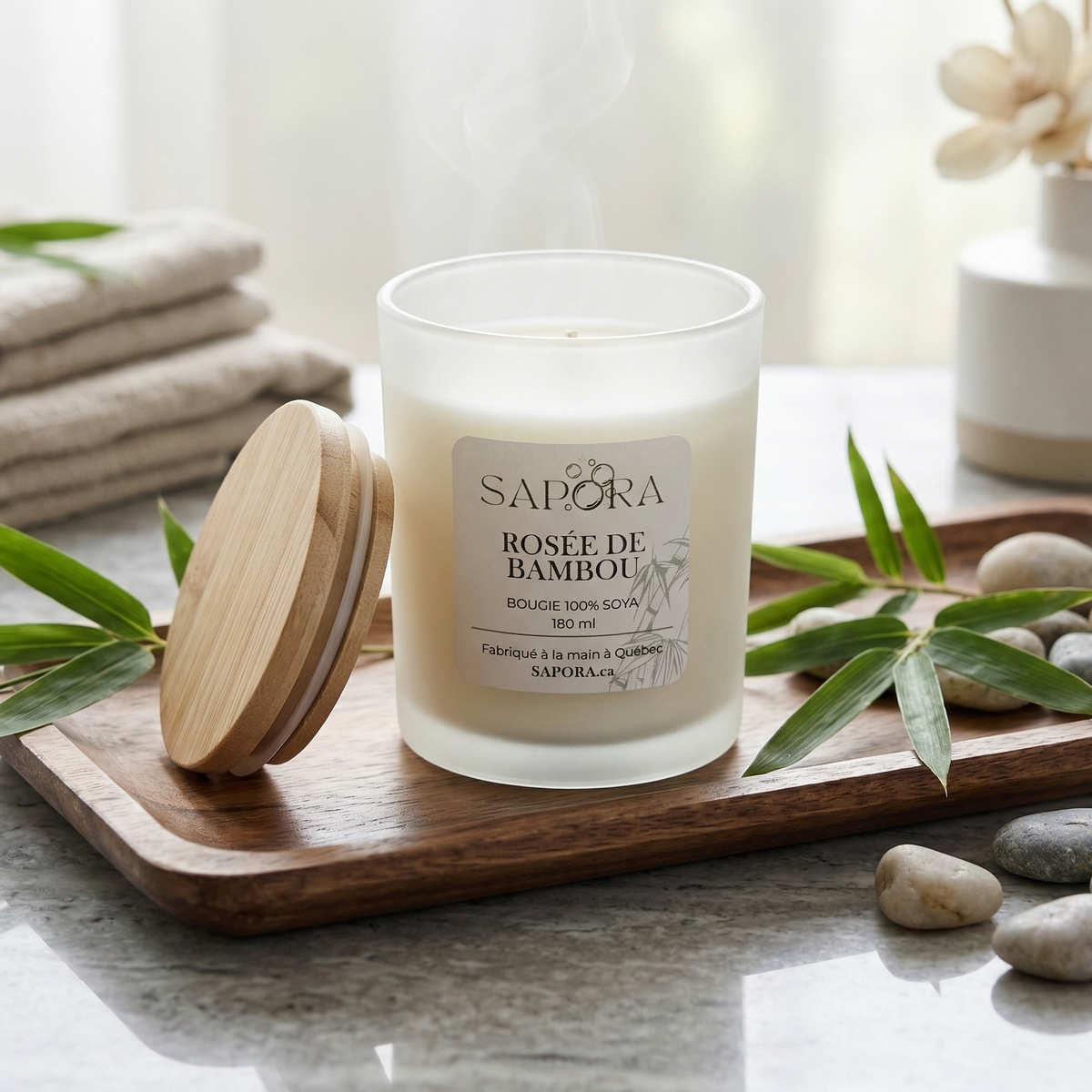 Bougie de soya artisanale Sapora Rosée de Bambou dans un pot en verre givré, posée sur un plateau de bois avec des feuilles de bambou et des pierres de rivière, ambiance spa zen et minimaliste.