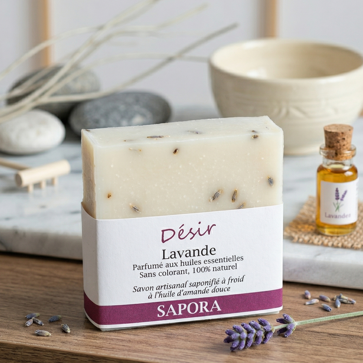 Savon artisanal saponifié à froid à l'huile d'amande douce Désir Lavande de Sapora, dans un décor zen avec pierres de massage, fiole d'huile essentielle et fleurs de lavande sur un plateau en marbre.