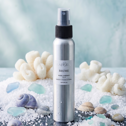 Bouteille de brume d'ambiance SAPORA en aluminium posée sur une pierre de schiste noire, entourée de gros sel marin, de verre de mer turquoise, de coquillages mauves et de corail blanc sur fond bleu clair.