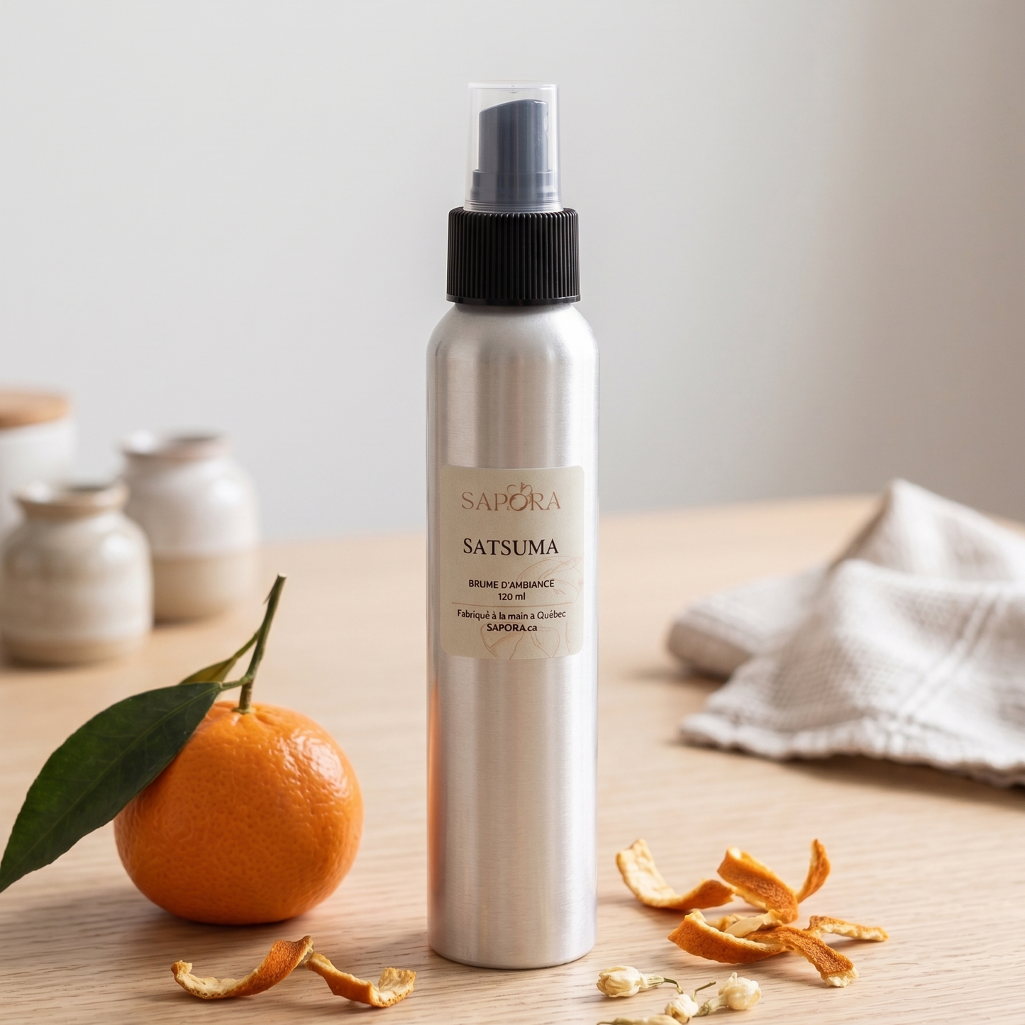 Flacon de bruine d'ambiance Sapora Satsuma en aluminium, posé sur une table en bois clair avec une mandarine fraîche, des zestes séchés et un décor zen minimaliste.
