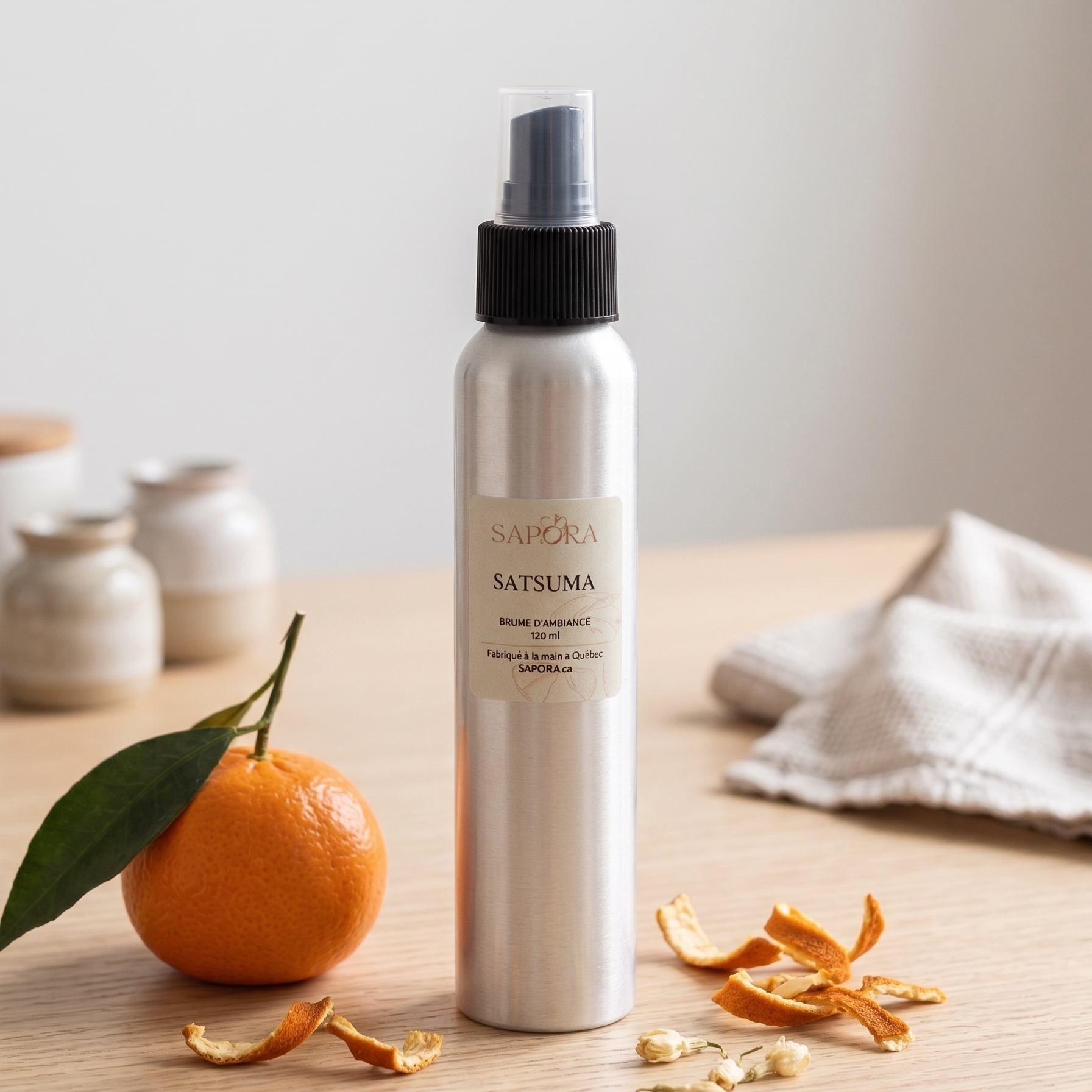 Flacon de bruine d'ambiance Sapora Satsuma en aluminium, posé sur une table en bois clair avec une mandarine fraîche, des zestes séchés et un décor zen minimaliste.