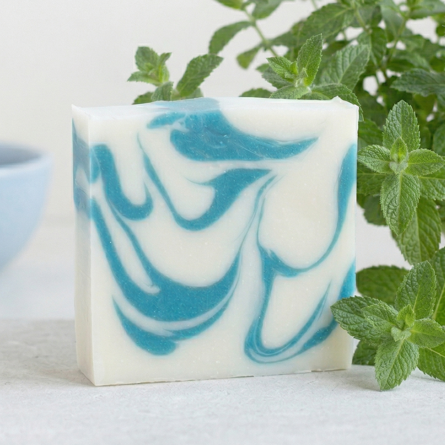 Savon artisanal blanc avec marbrures bleu turquoise, sans étiquette, posé sur une pierre claire. En arrière-plan, un plant de menthe fraîche et un bol bleu minimaliste sur fond blanc épuré.