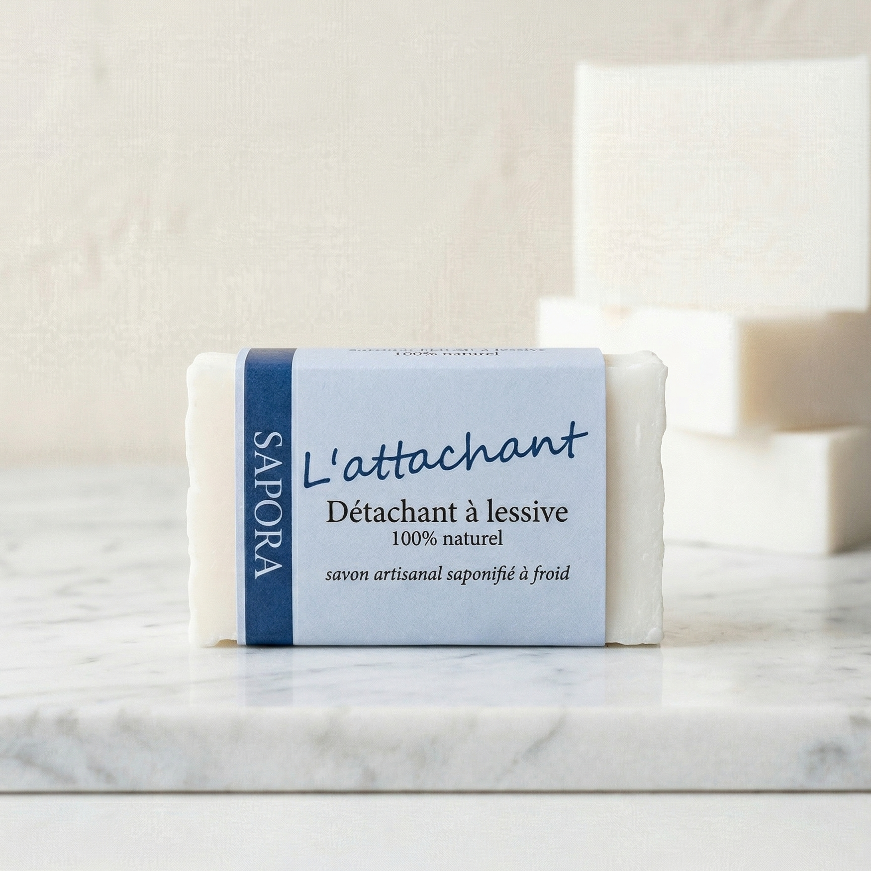 Savon détachant à lessive L'attachant de Sapora, savon artisanal blanc saponifié à froid présenté sur un socle en marbre blanc dans un décor minimaliste et épuré.