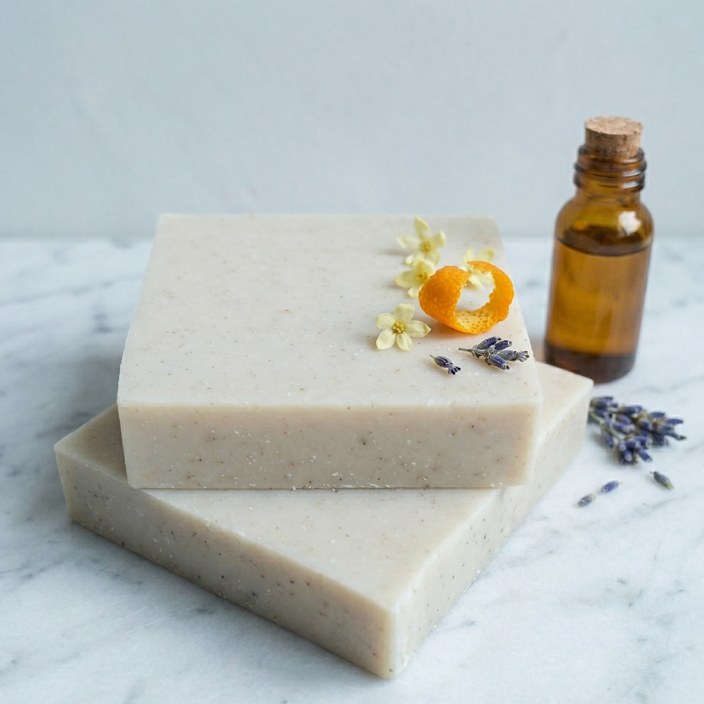 Savon artisanal Harmonie aux huiles essentielles d'Ylang-Ylang, orange et lavande, posé sur un marbre blanc avec des fleurs d'oranger et une fiole d'apothicaire.