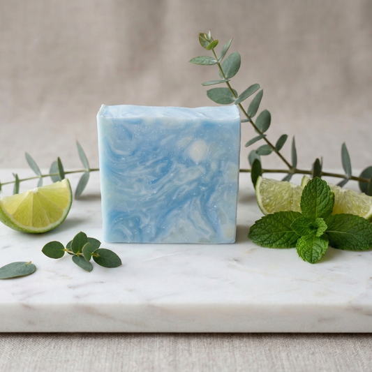 Savon artisanal "PUR" de Sapora aux marbrures bleu clair et blanc, posé sur une planche de marbre. Décor épuré avec des quartiers de lime fraîche, des feuilles de menthe et des branches d'eucalyptus. Ambiance lumineuse et rafraîchissante.