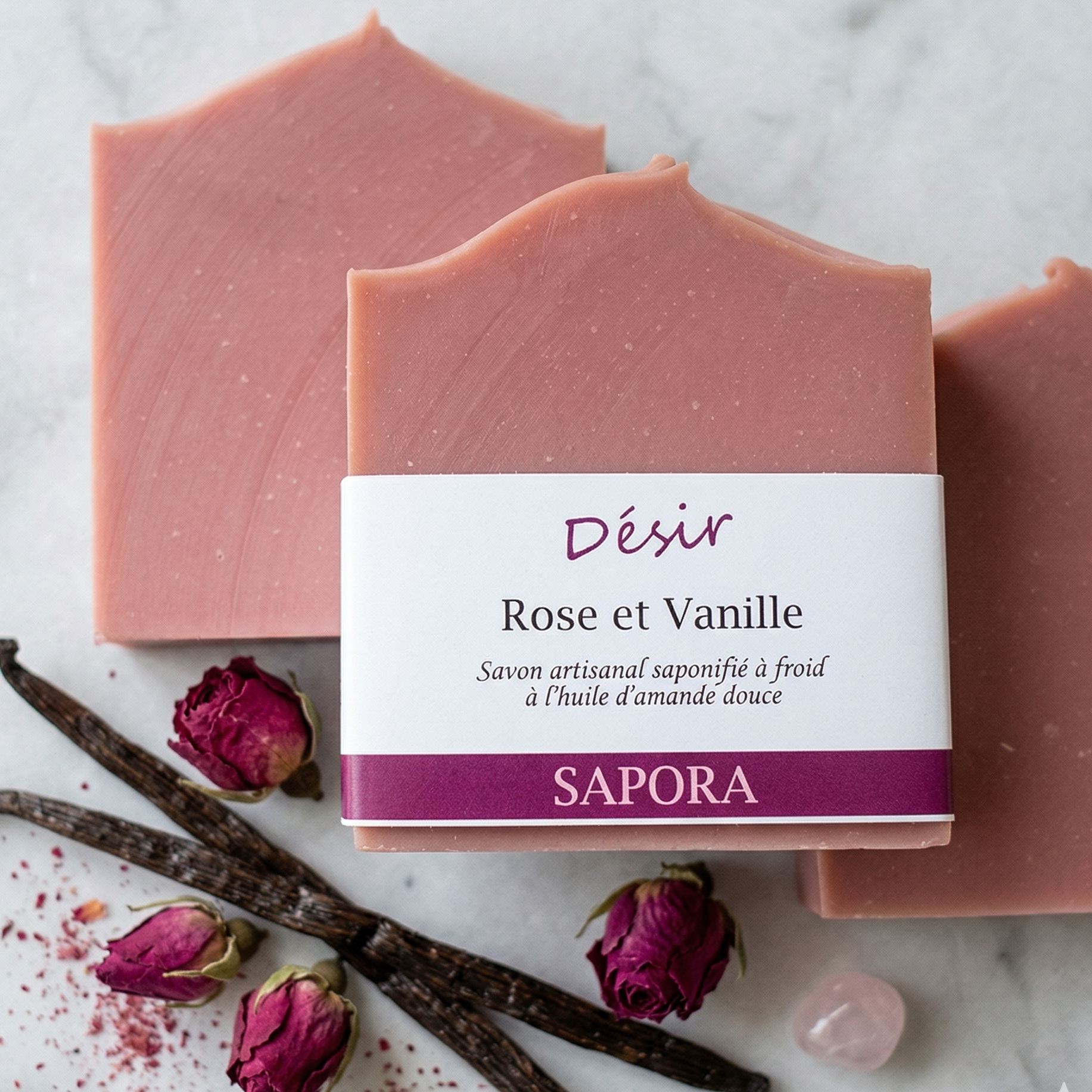 Savon artisanal rose 'Désir' de Sapora avec étiquette, posé sur un fond de marbre blanc épuré. Accompagné de gousses de vanille naturelle, de boutons de rose séchés et d'un cristal de quartz rose.
